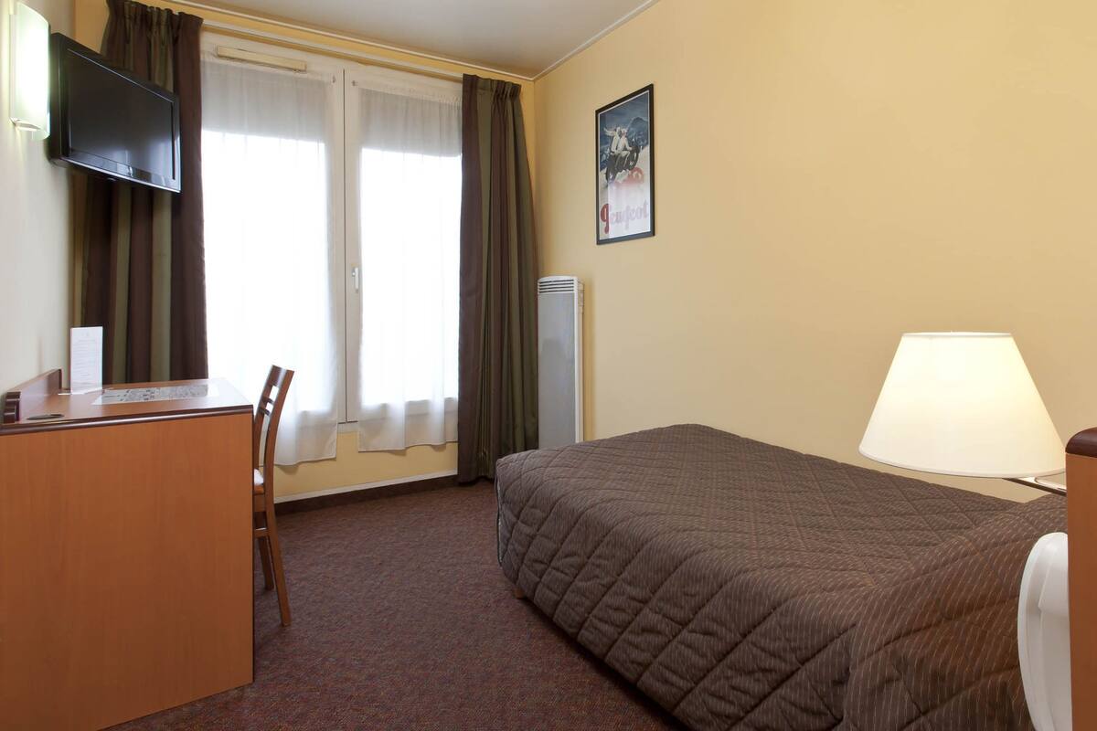Hotellihuone, jossa on sänky, työpöytä, tuoli, televisio, lamppu ja juliste seinällä.