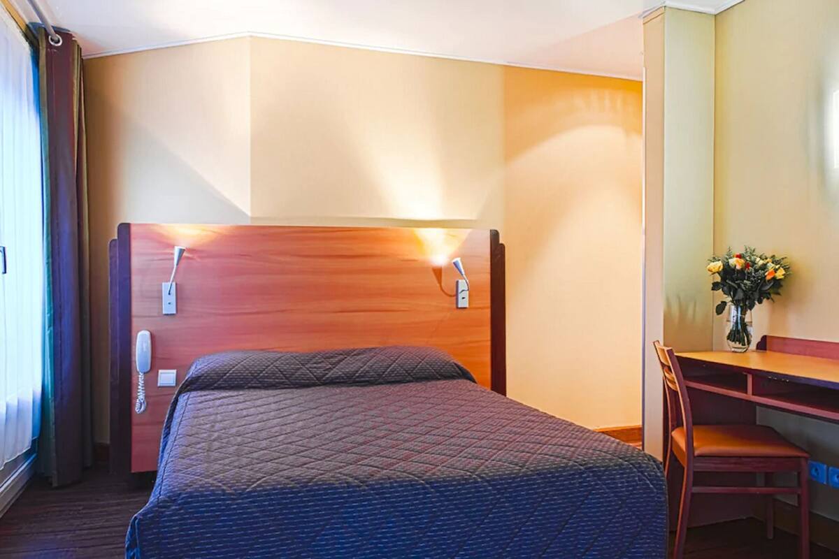Hotellihuone, jossa on sänky, työpöytä, tuoli, puhelin ja maljakko, jossa on kukkia.