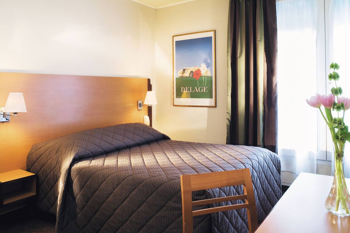 Hotellihuone, jossa on sänky, yöpöytä, lamppu, seinällä oleva kehystetty kuva, ikkuna verhoilla ja pöydällä oleva maljakko, jossa on kukkia.