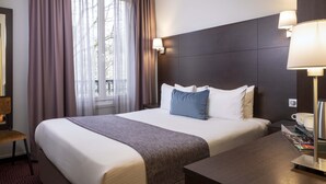 A hotel room with a large bed, a wooden nightstand, a desk, and a window with curtains.