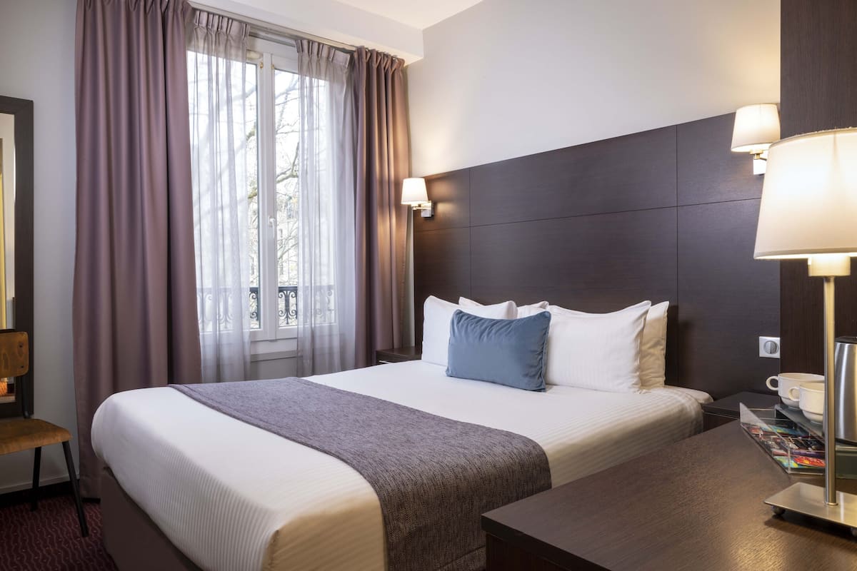 Une chambre d’hôtel avec un grand lit, une table de chevet en bois, un bureau et une fenêtre avec des rideaux.