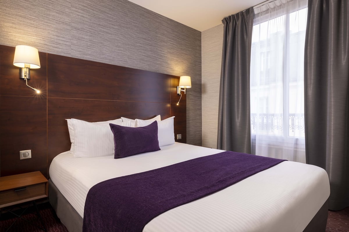 Une chambre d’hôtel avec un grand lit, deux lampes fixées au mur, une fenêtre avec des rideaux et une table de chevet.