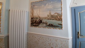 Um quarto com radiador fixado na parede, porta com puxador dourado e quadro emoldurado representando uma cena portuária.