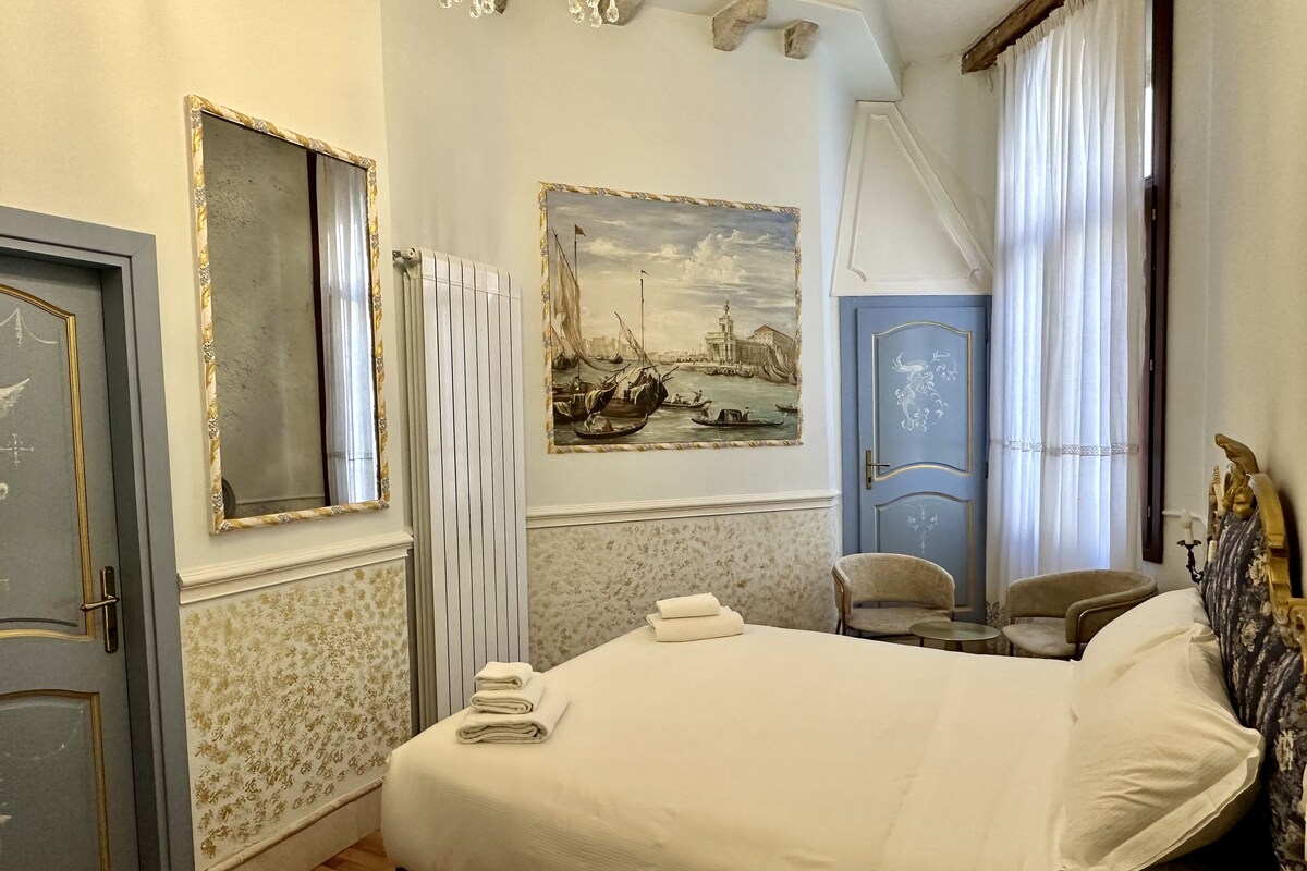 Un letto rifatto con asciugamani piegati, un quadro raffigurante una scena storica, una porta blu e una sedia.