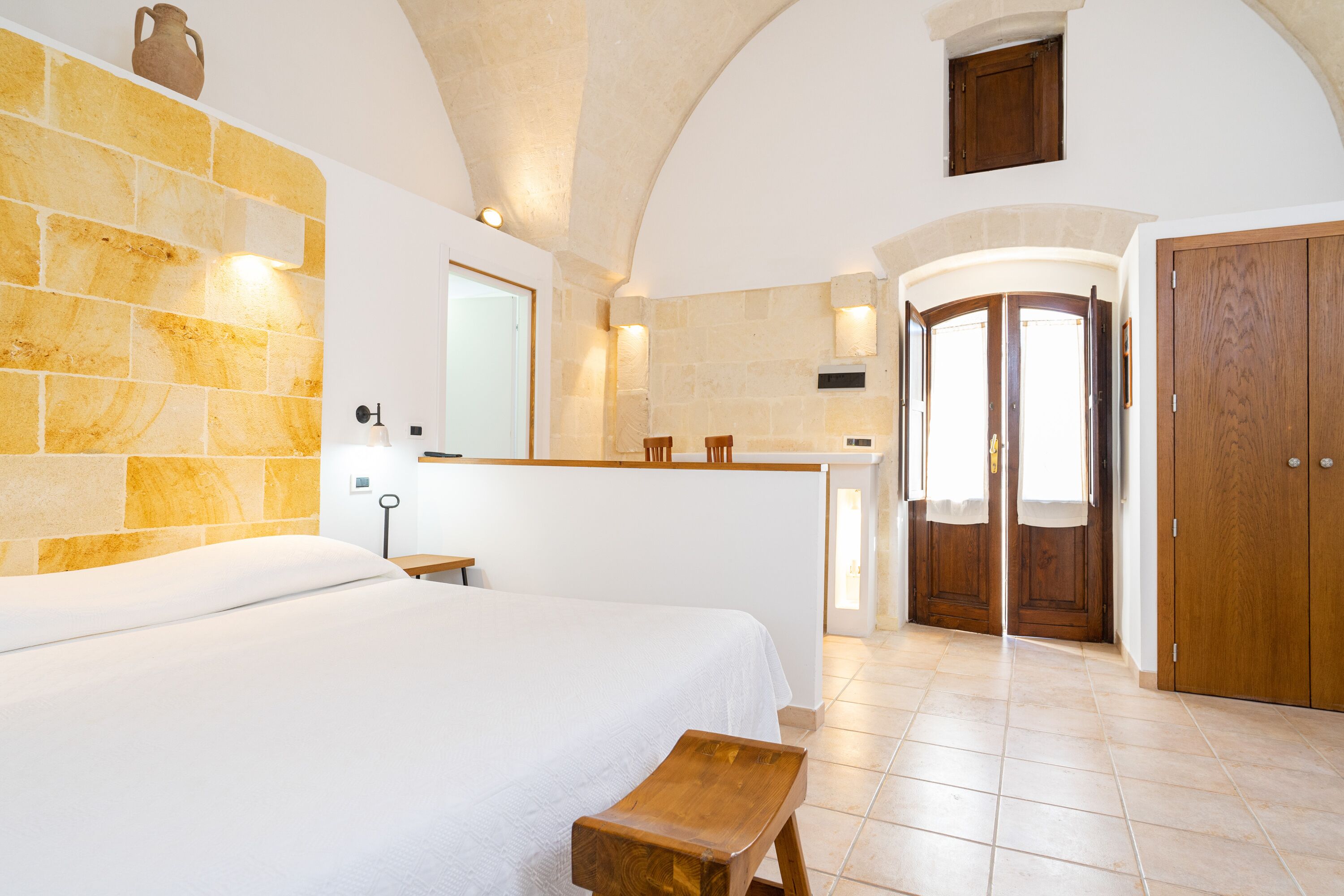 A bedroom with a large bed, a wooden bench, a stone wall, and a kitchen area with a wooden door.