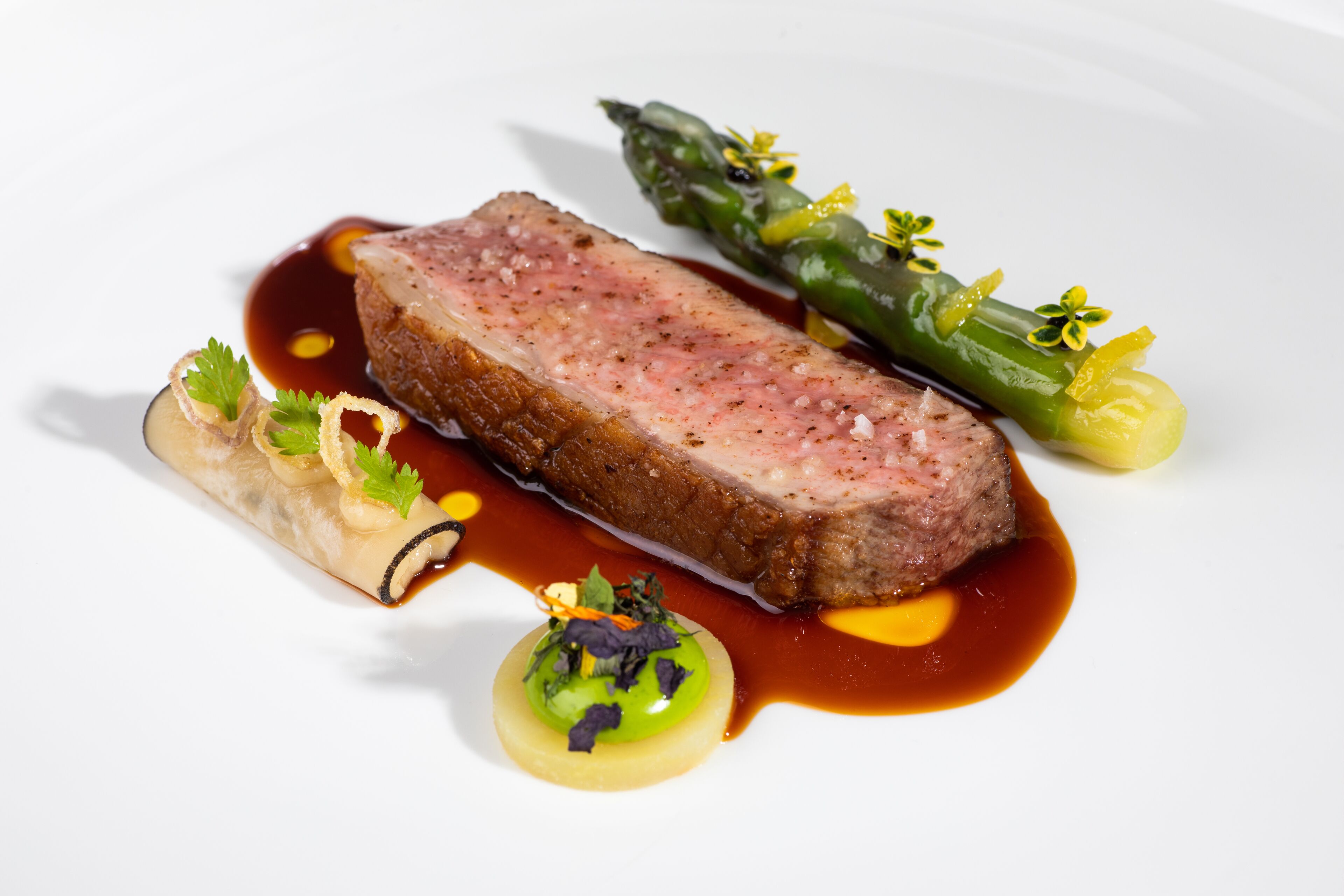 Un plato con una porción de carne, espárragos y un recipiente pequeño de salsa.
