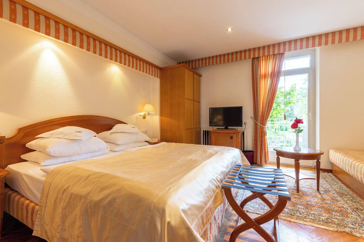 A hotel room with a large bed, a TV, a small table, and a window with curtains.