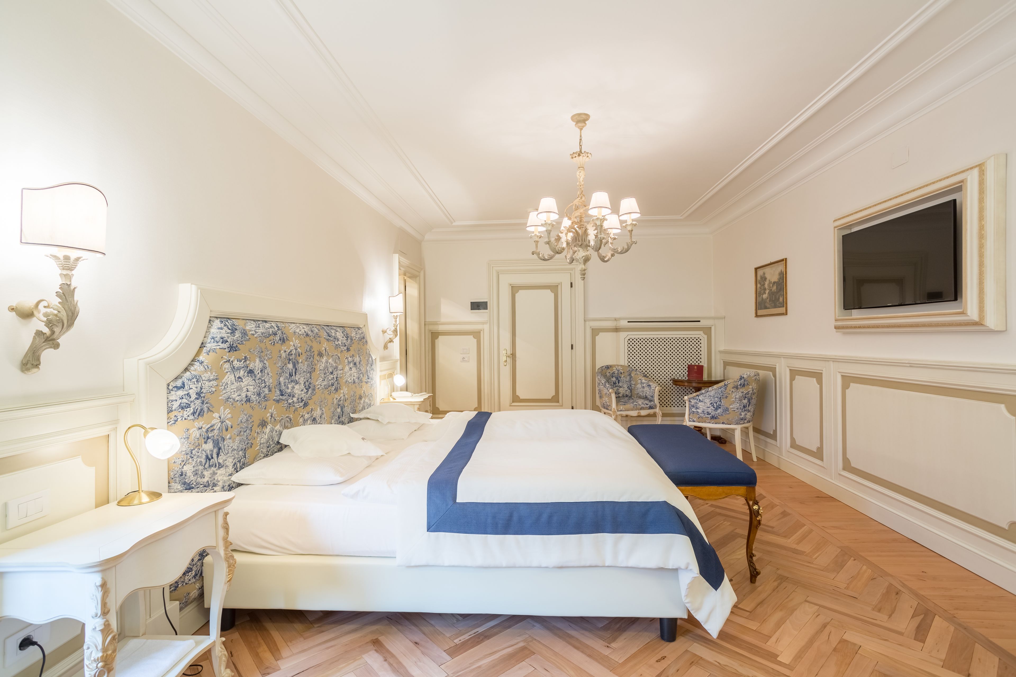 A hotel room with a large bed, a bedside table with a lamp, a chair, a TV, and a chandelier.