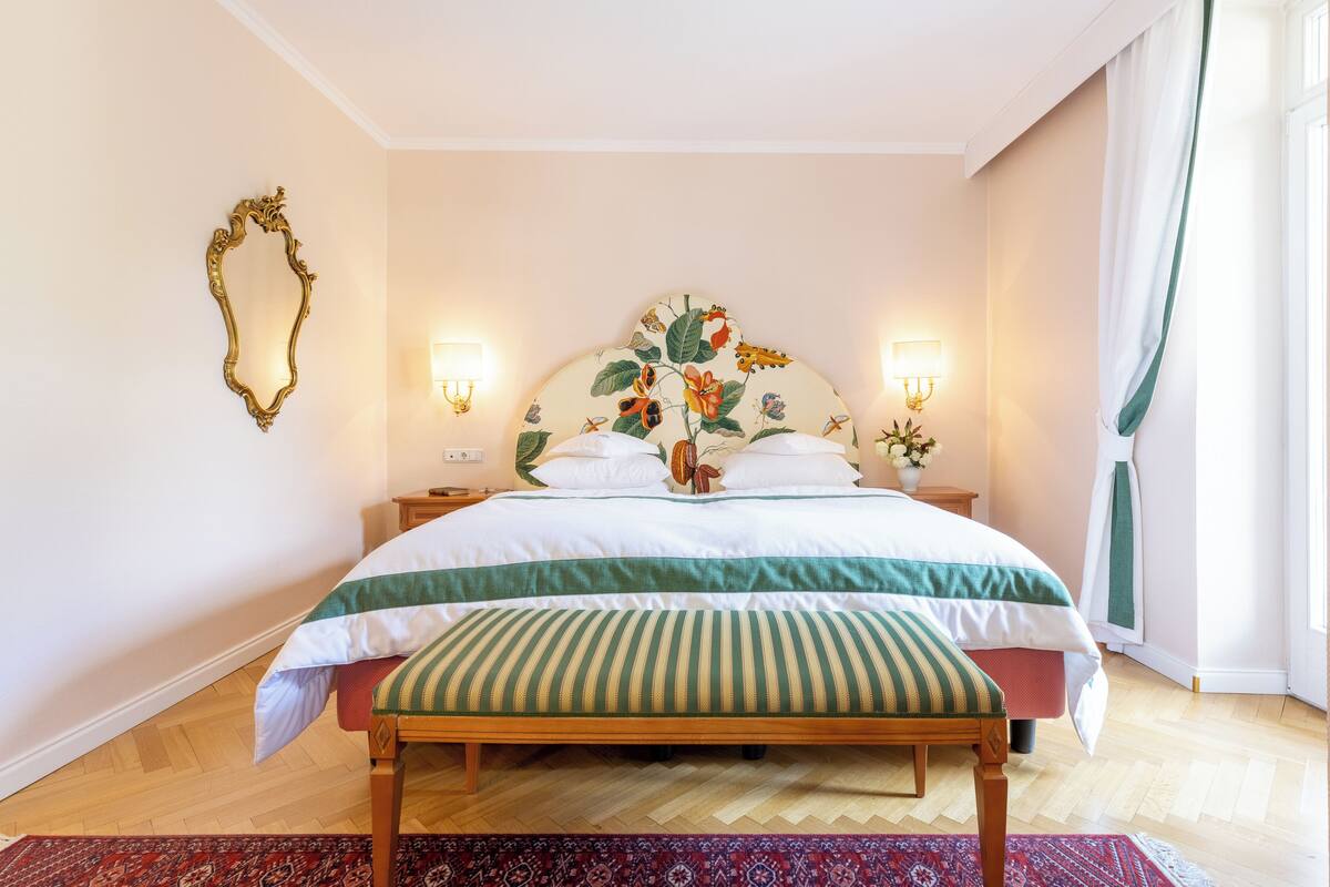 A bedroom with a large bed, a wooden bench, a floral headboard, two wall sconces, and a patterned rug.