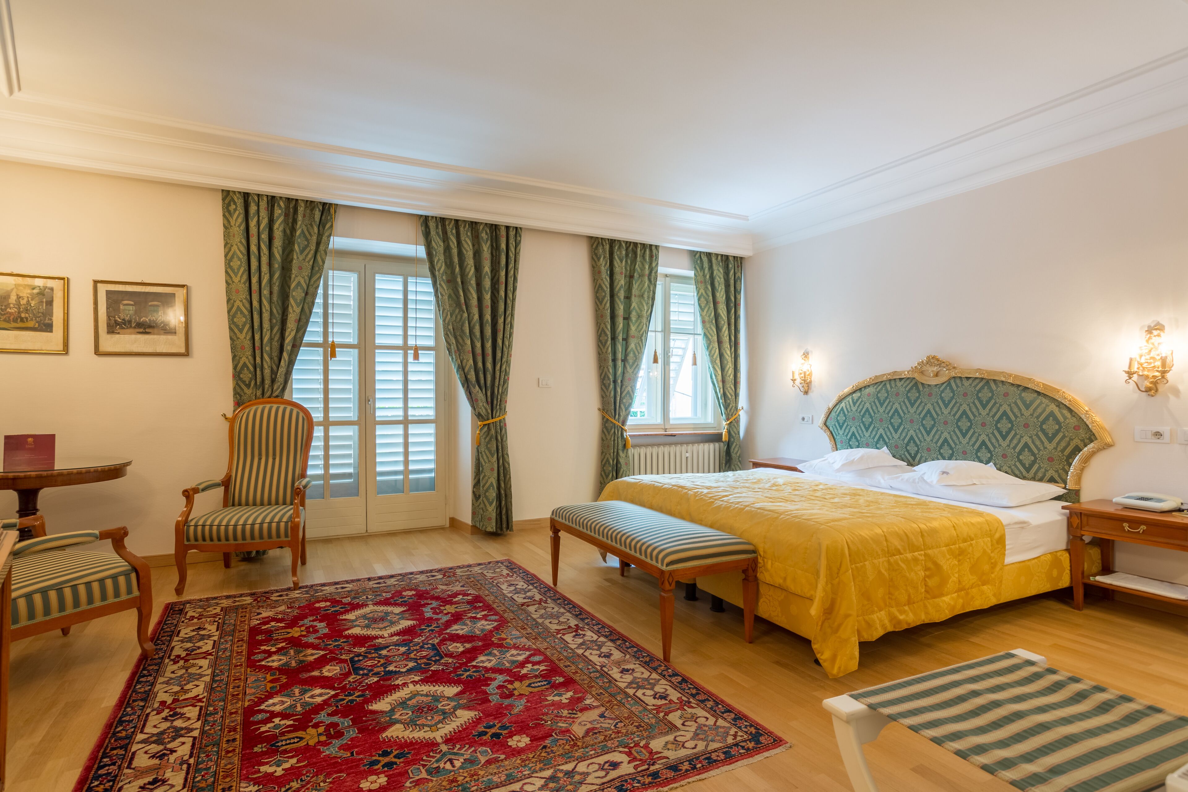 A hotel room with a bed, a bench, a chair, a table, and a large patterned rug.