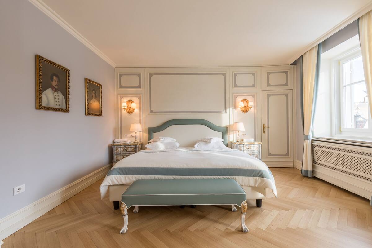 A neatly arranged bedroom with a large bed, two bedside tables with lamps, a bench, and two framed pictures on the wall.