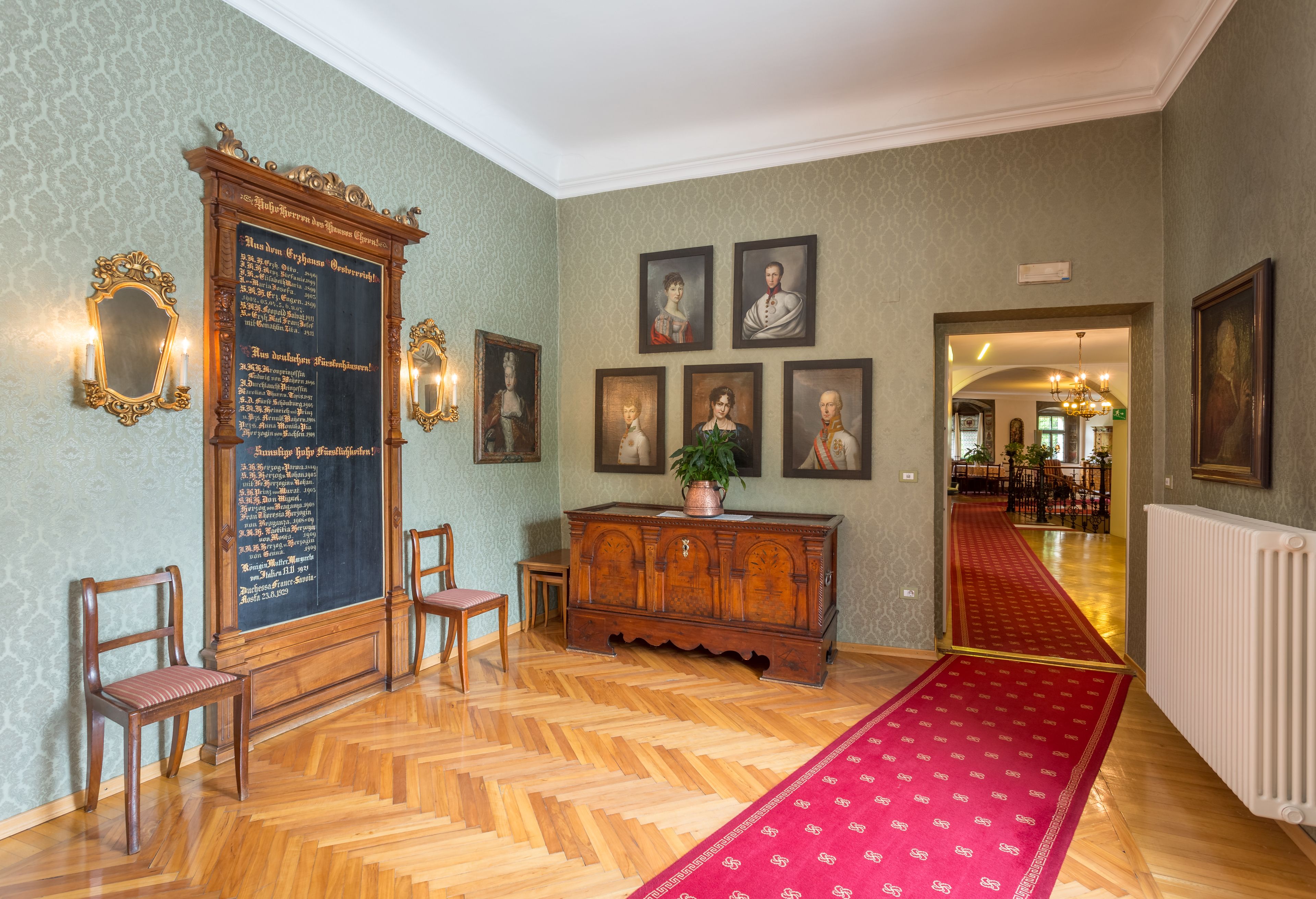 A room with a wooden floor, a red carpet, a wooden cabinet, and framed portraits on the wall.