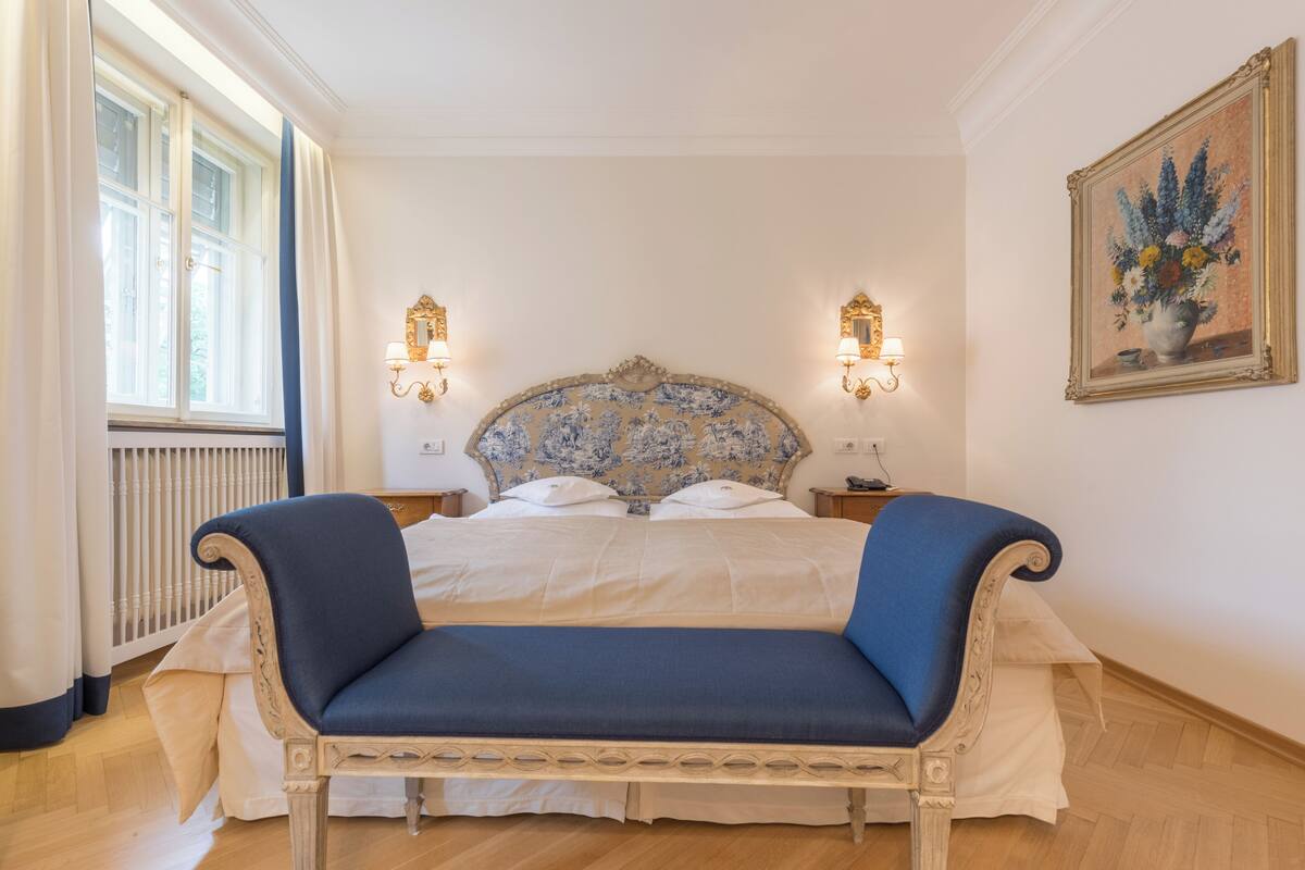 A bedroom with a large bed, two bedside tables, a nightstand, a window with curtains, a wall-mounted painting, and a wooden floor.
