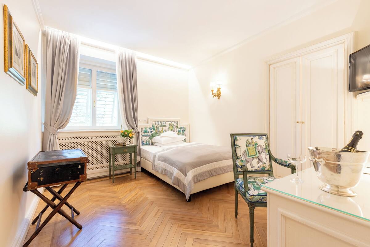 A bedroom with a bed, a desk, a chair, a nightstand, a window with curtains, and a television on the wall.