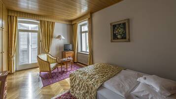 Ein Schlafzimmer mit Bett, Stuhl, kleinem Tisch, Fernseher und einem Fenster mit Vorhängen.