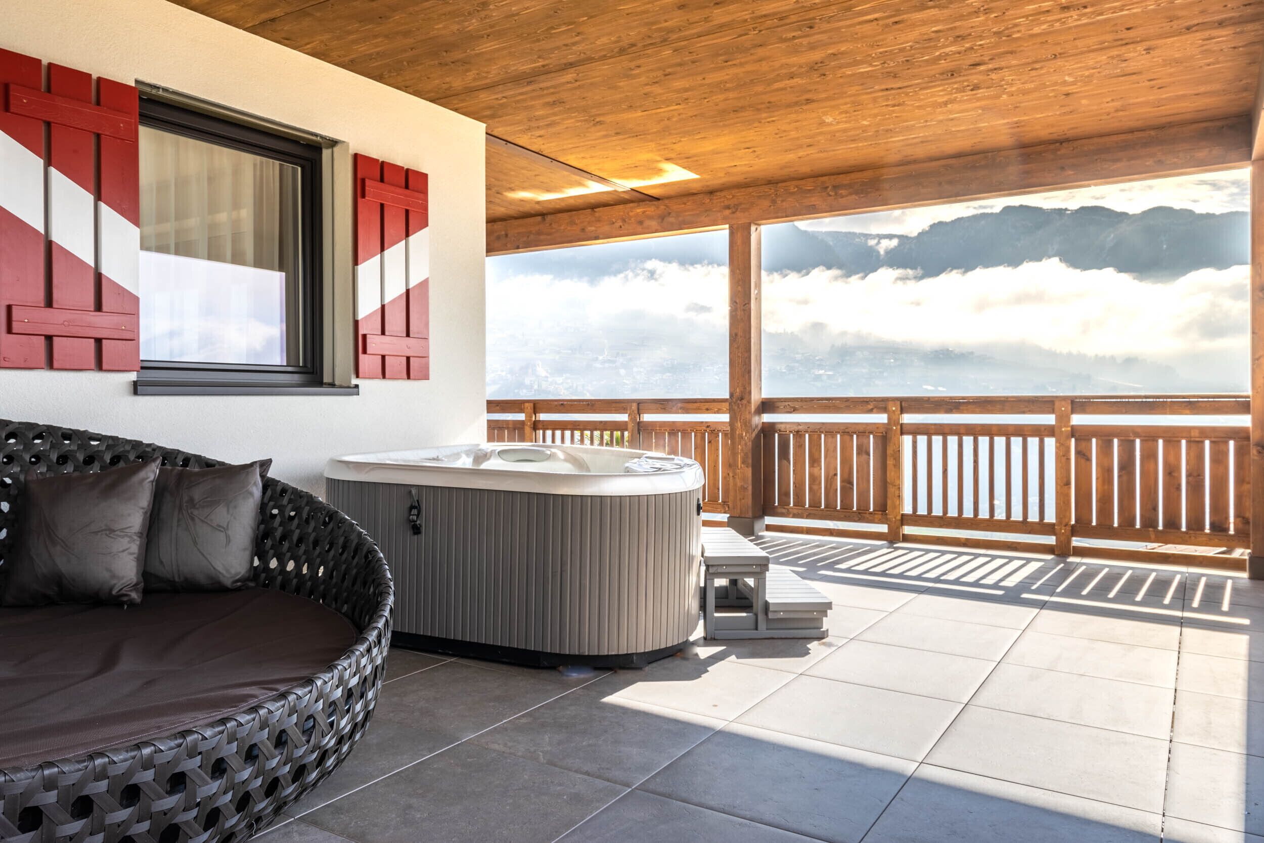 A hot tub and a wicker sofa on a balcony with a wooden railing, overlooking a mountainous landscape.