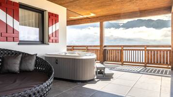 A hot tub and a wicker sofa on a balcony with a wooden railing, overlooking a mountainous landscape.