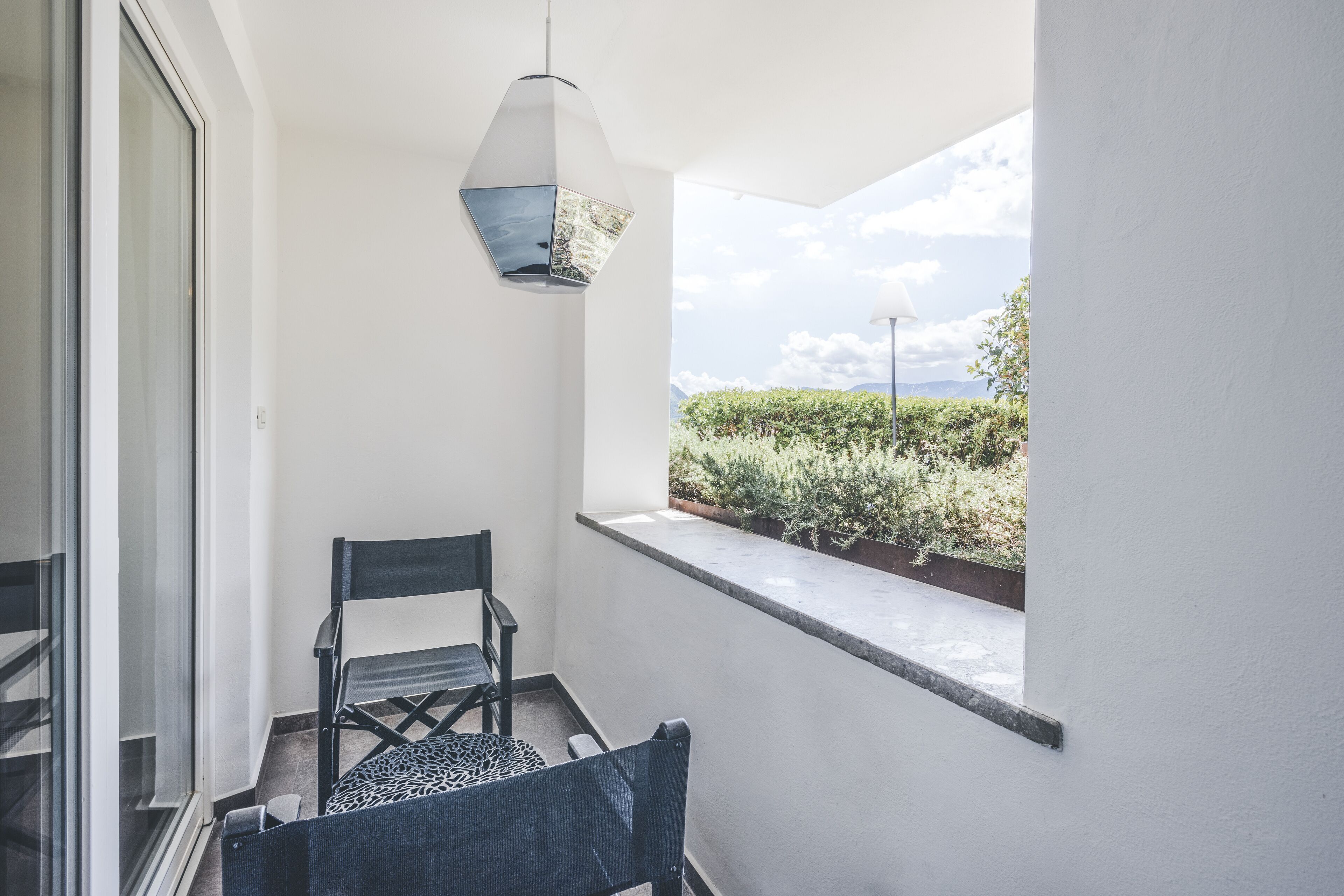 A balcony with a view of greenery, a chair, and a hanging light fixture.