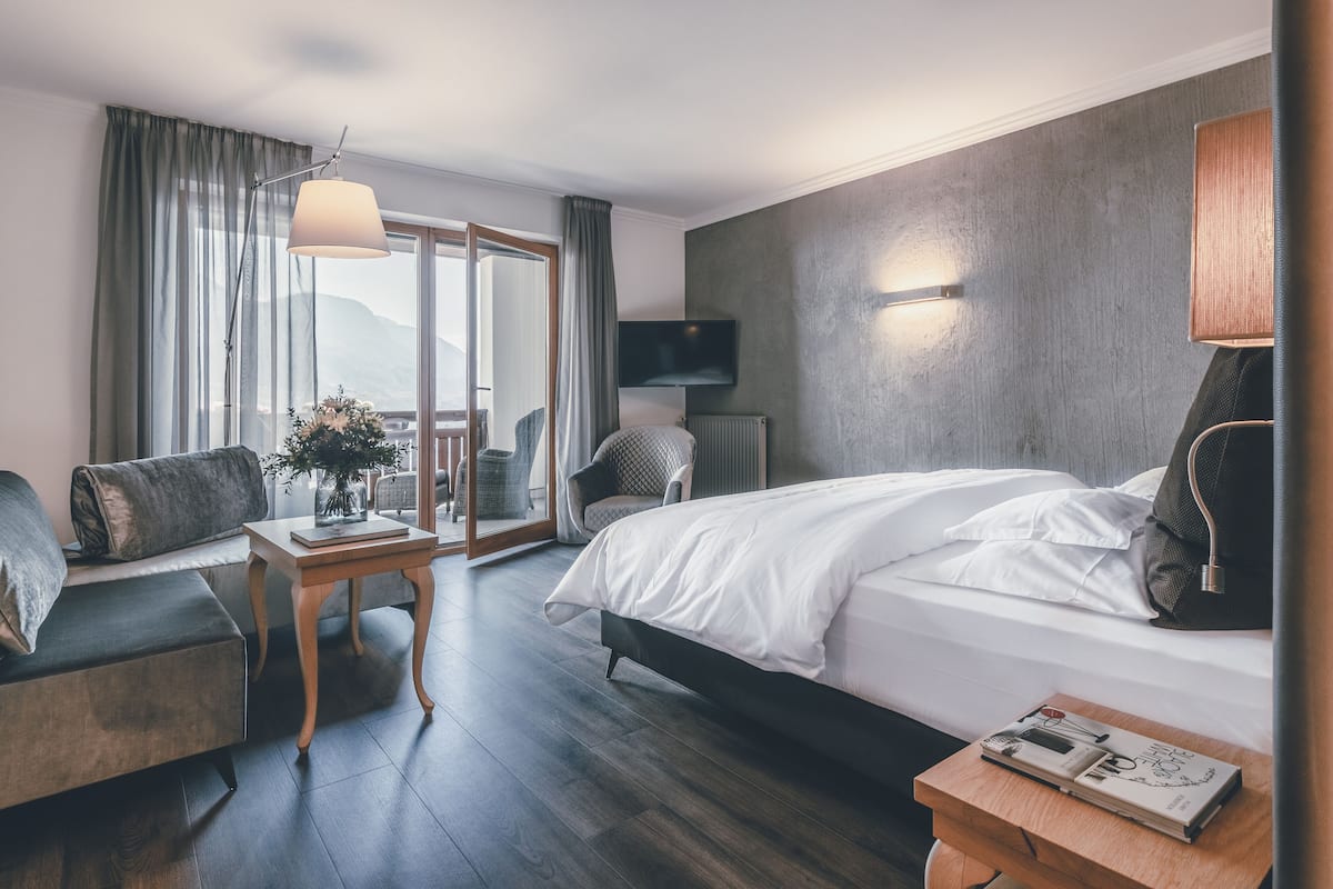 A modern hotel room with a large bed, a sofa, a small table with a vase, and a view of the outdoors.