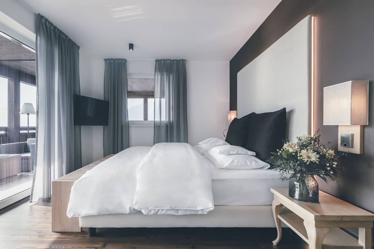 A modern hotel room with a large bed, a bedside table with a vase of flowers, a flat-screen TV, and a large window with curtains.