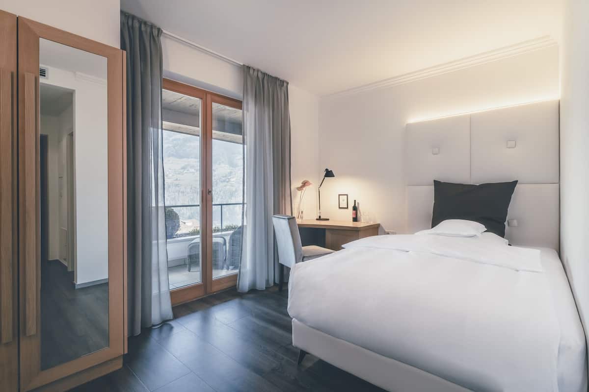 A hotel room with a bed, a desk, a chair, a lamp, and a view of the outdoors.