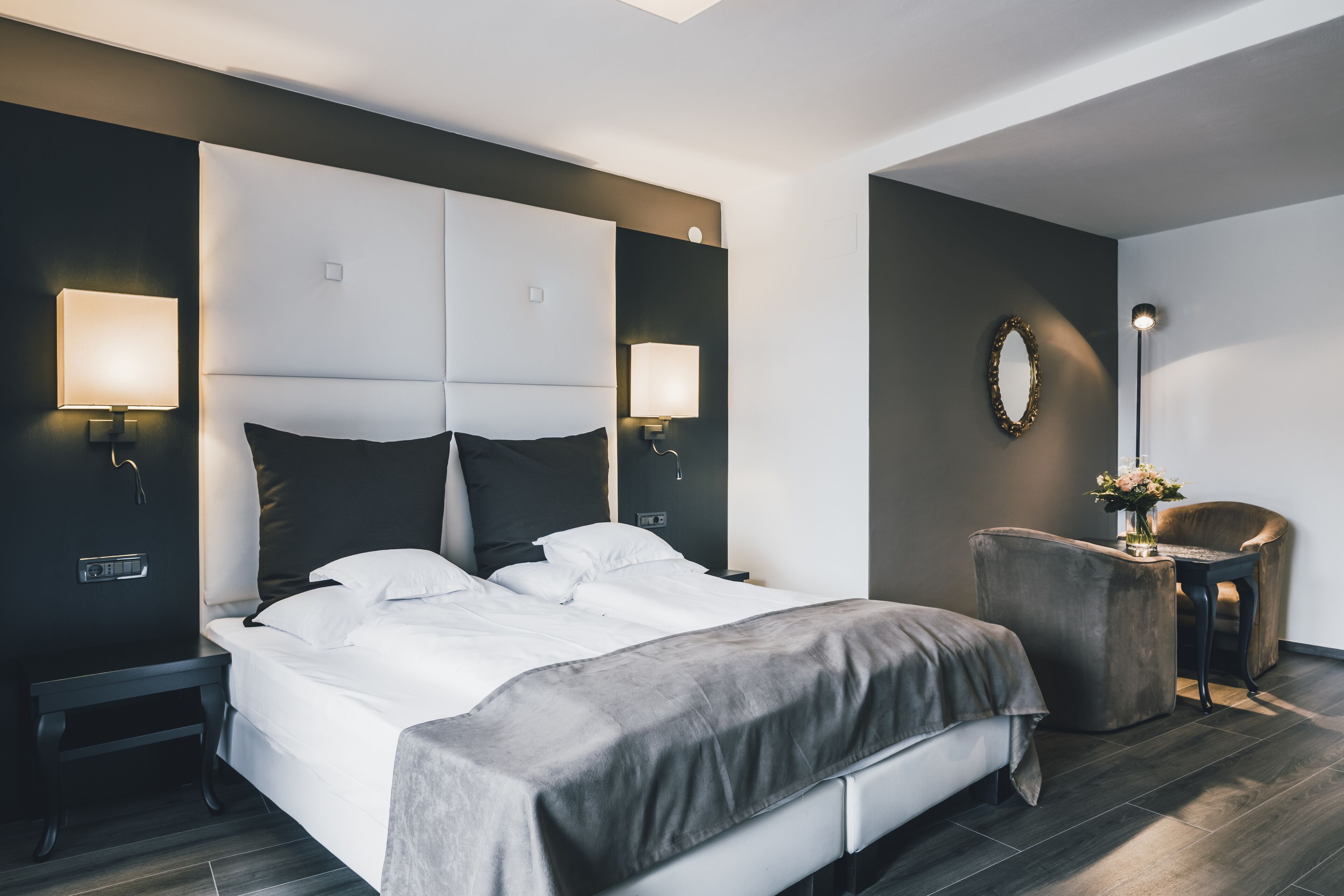A modern hotel room with a large bed, bedside lamps, a small table with a vase, and a chair.
