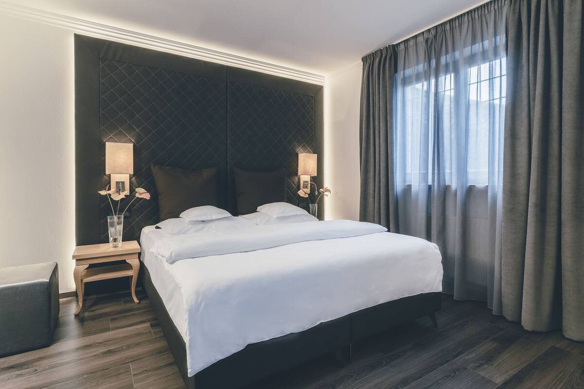 A hotel room with a large bed, two bedside tables with lamps, a dark headboard, and a view of the outdoors through sheer curtains.