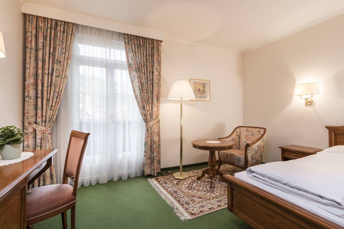 A hotel room with a bed, a desk with a lamp, a chair, a small table, and a floor lamp.