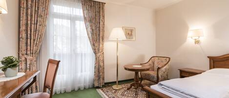 Una habitación de hotel con una cama, un escritorio con lámpara, una silla, una mesita y una lámpara de pie.