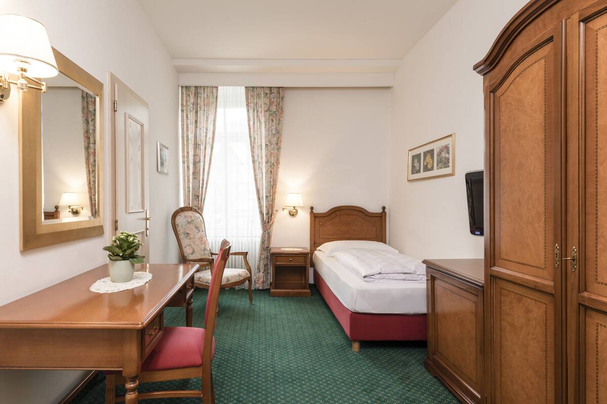 A hotel room with a wooden desk, chair, bed, wardrobe, and a mirror.