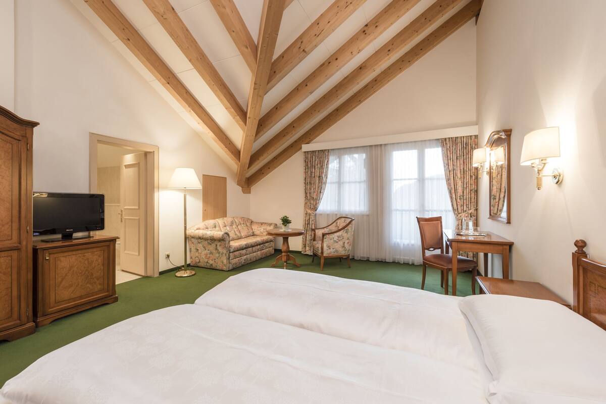 A hotel room with a large bed, a sofa, a chair, a small table, a TV, and a wooden ceiling.