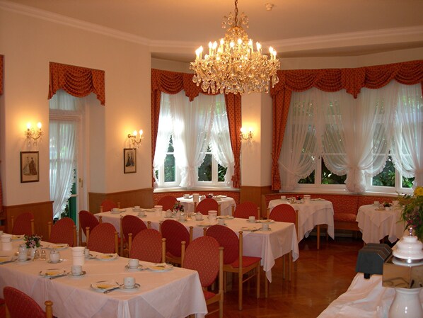 A dining room with tables set for a meal, chairs, a chandelier, and windows with curtains.