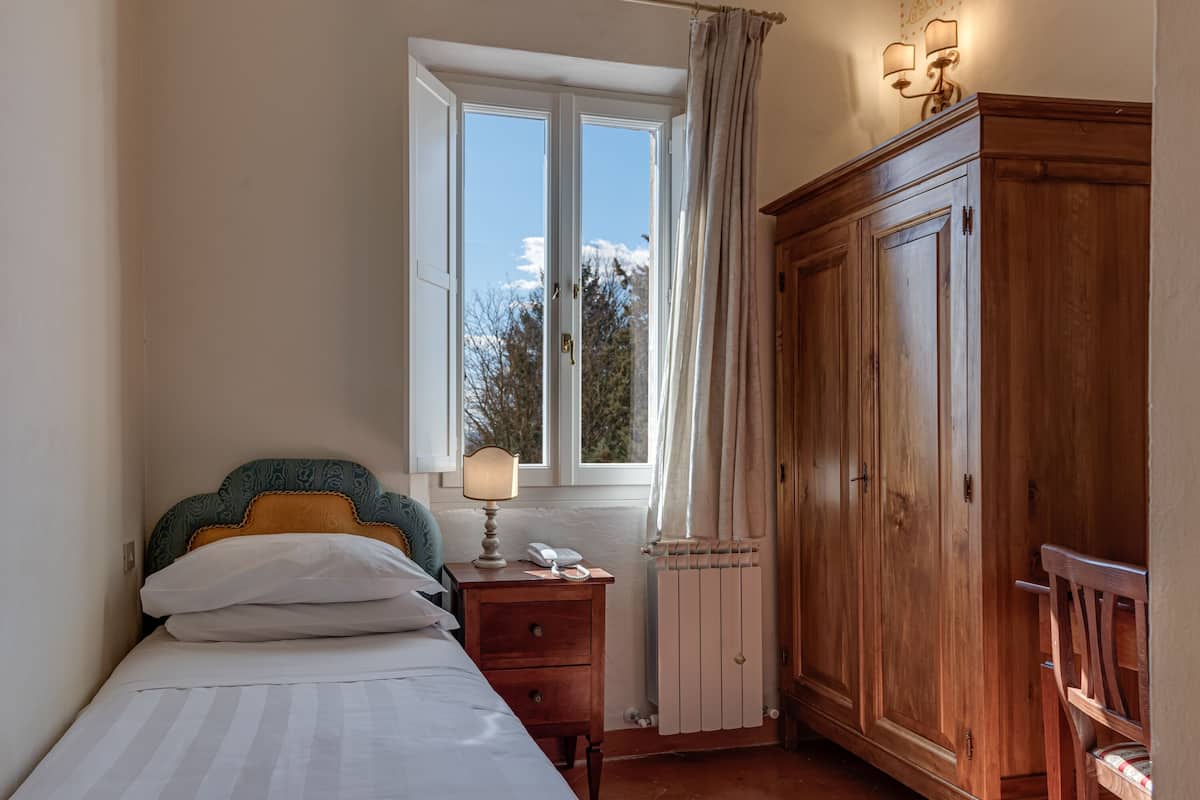 A bedroom with a bed, a nightstand, a wooden wardrobe, a window with curtains, and a radiator.