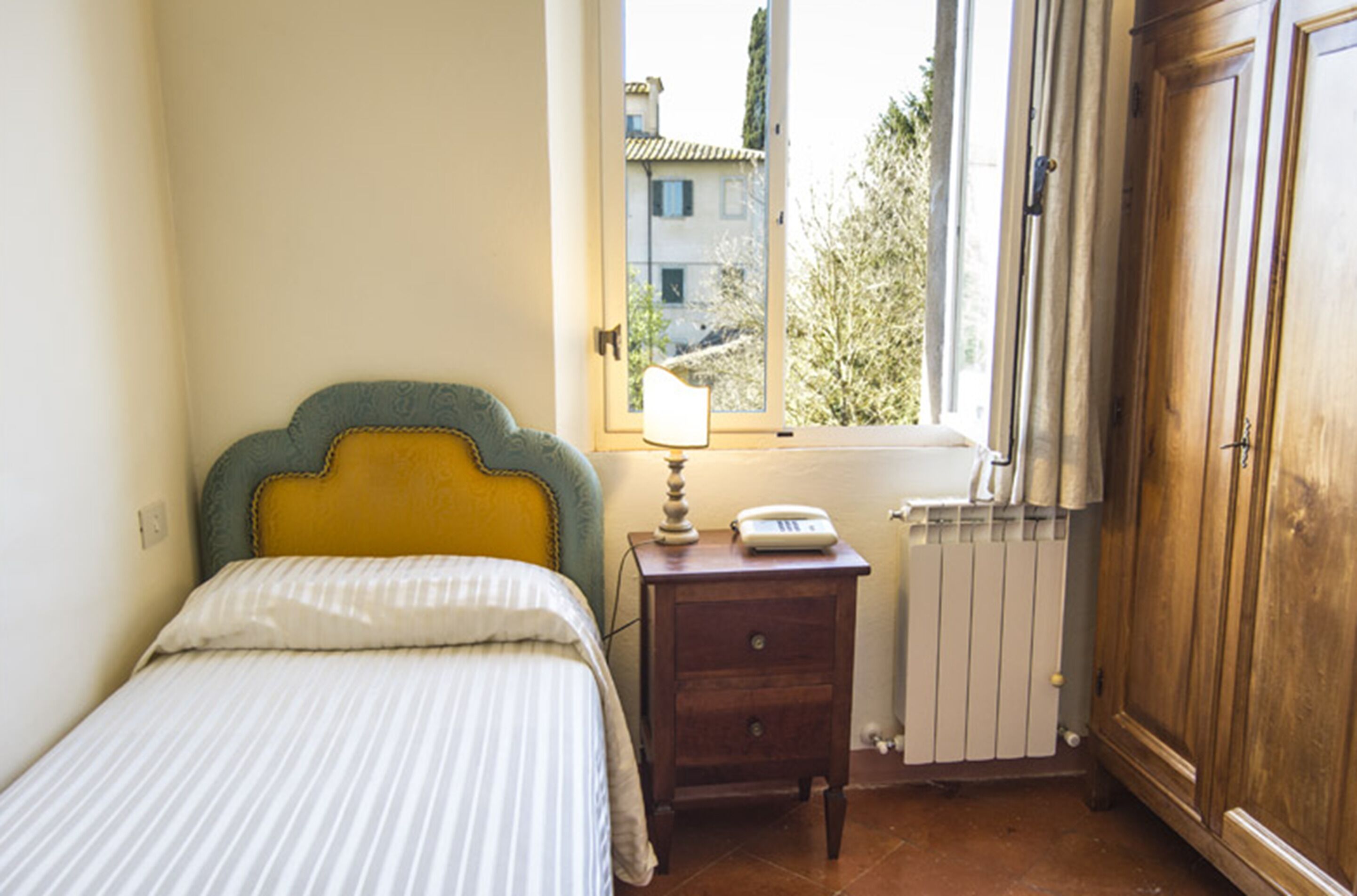 A bedroom with a bed, a bedside table with a lamp, a radiator, and a window with a view of buildings and trees.
