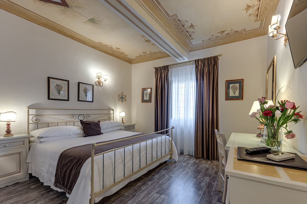 A hotel room with a bed, bedside tables, a desk with a chair, a mirror, and a window with curtains.
