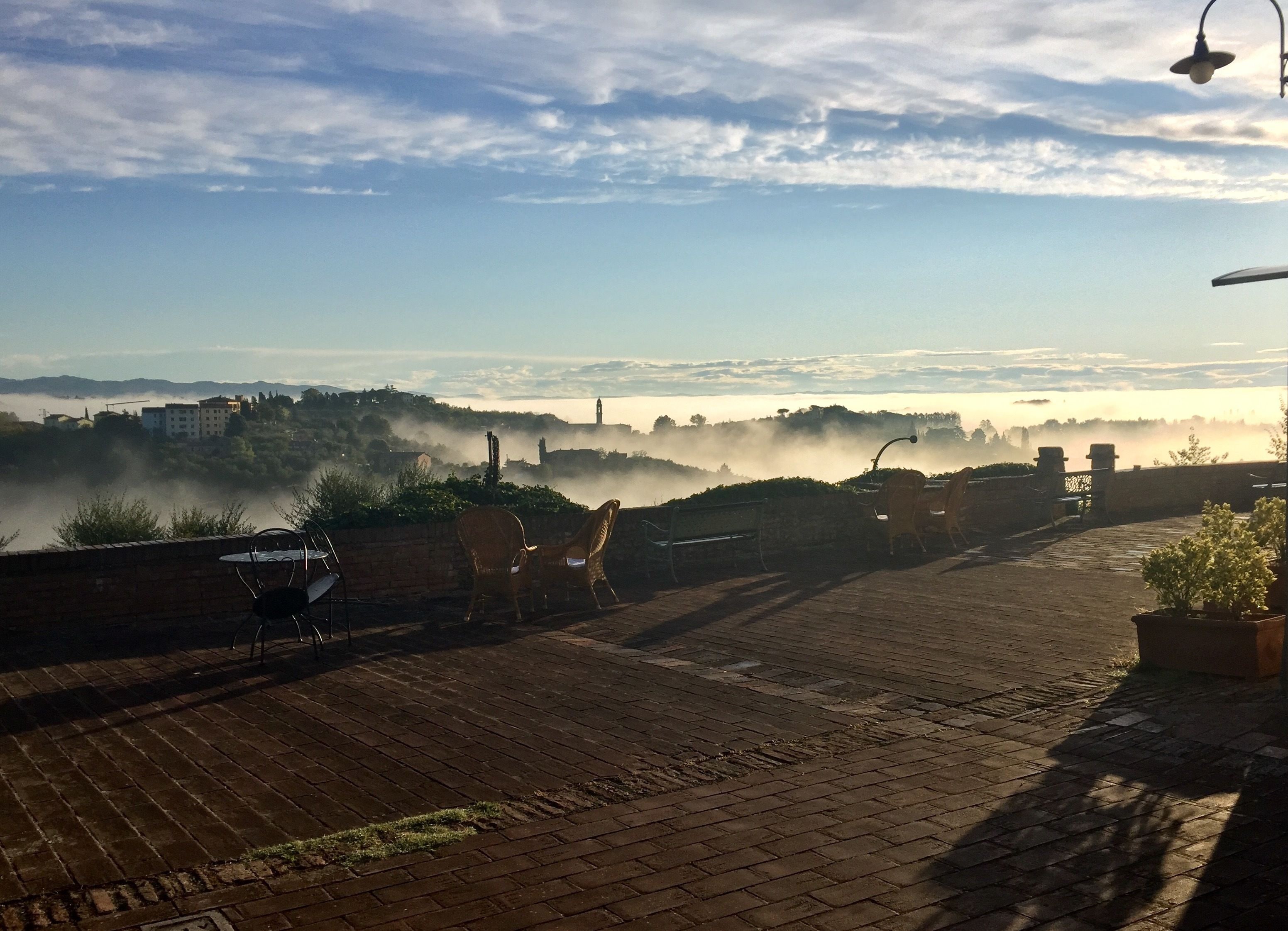Una terraza empedrada con sillas de mimbre y una planta en maceta, con vistas a un paisaje brumoso con edificios y una torre del reloj a lo lejos.