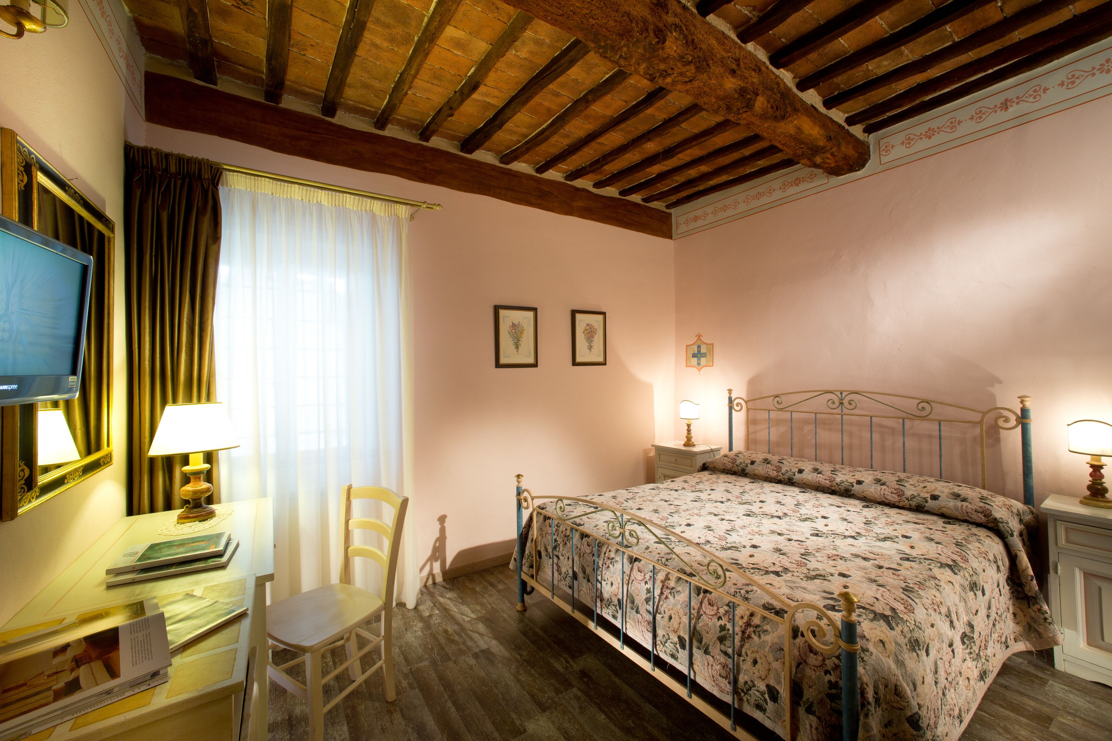 A bedroom with a wooden ceiling, a bed with a floral bedspread, a desk with a lamp, and a chair.