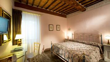A bedroom with a wooden ceiling, a bed with a floral bedspread, a desk with a lamp, and a chair.
