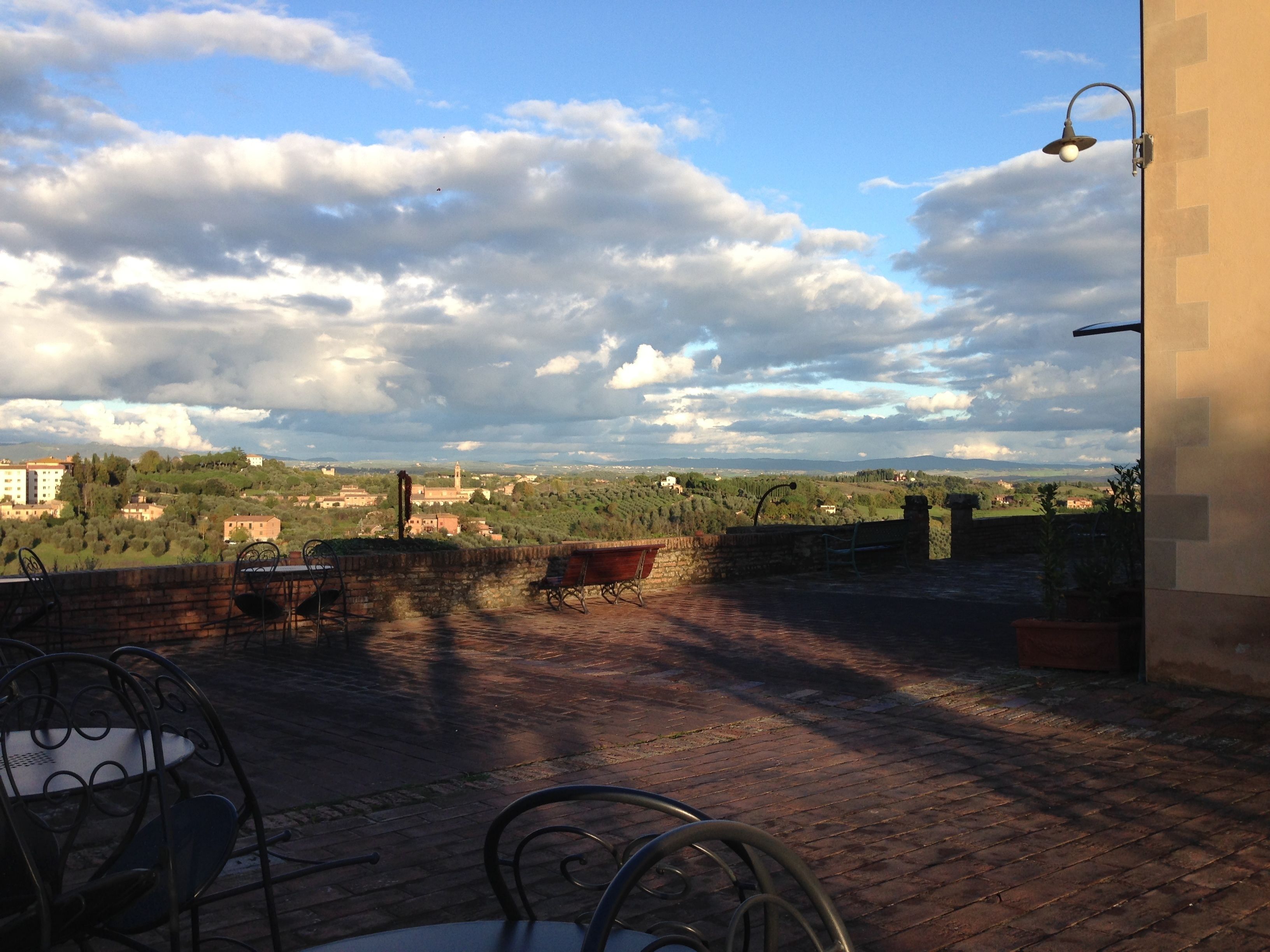 Una terraza con suelo de ladrillo, sillas de metal y una mesa. Una lámpara está fijada a la pared. La vista muestra un paisaje lejano con edificios y árboles.
