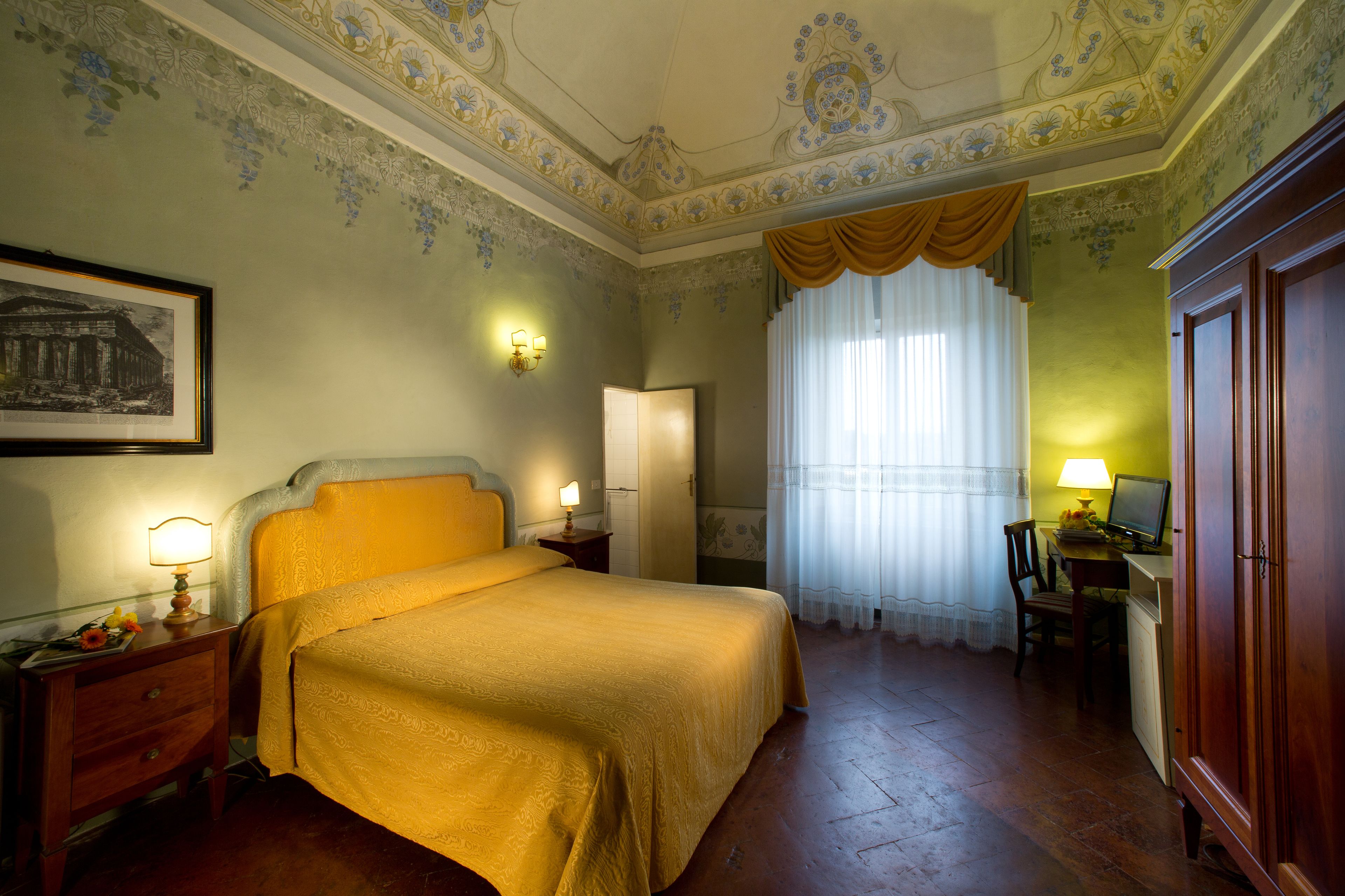 A hotel room with a bed, a desk with a computer, a chair, a nightstand with a lamp, and a framed picture on the wall.