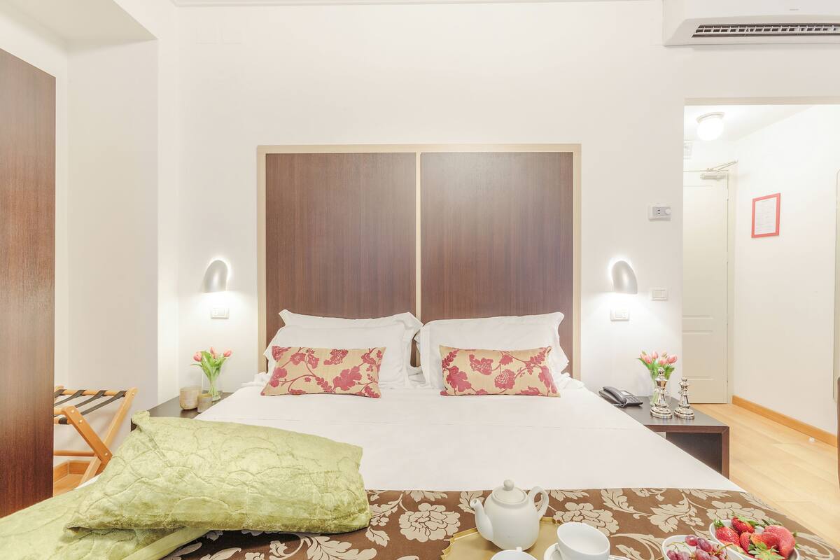 A hotel room with a double bed, two bedside lamps, a bedside table with a phone, a small vase with flowers, a green pillow, a brown and white patterned bedspread, a white teapot, and a tray with a cup and strawberries.