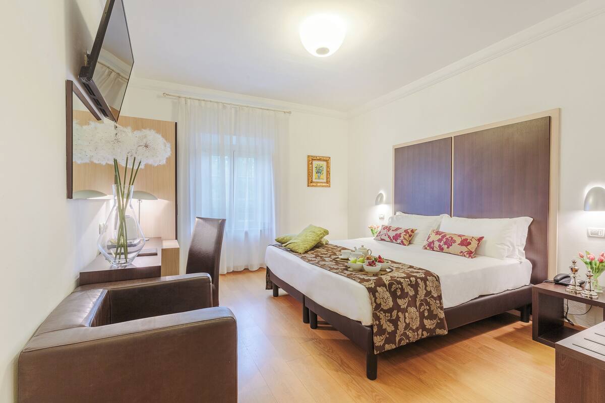 A hotel room with a bed, bedside table, lamp, chair, and a desk with a computer.