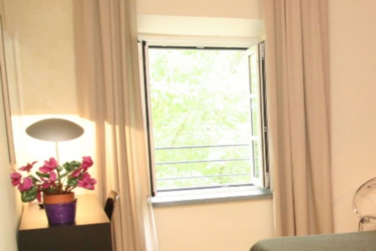 A bedroom with a bed, a window with curtains, a chair, and a wall-mounted light fixture.