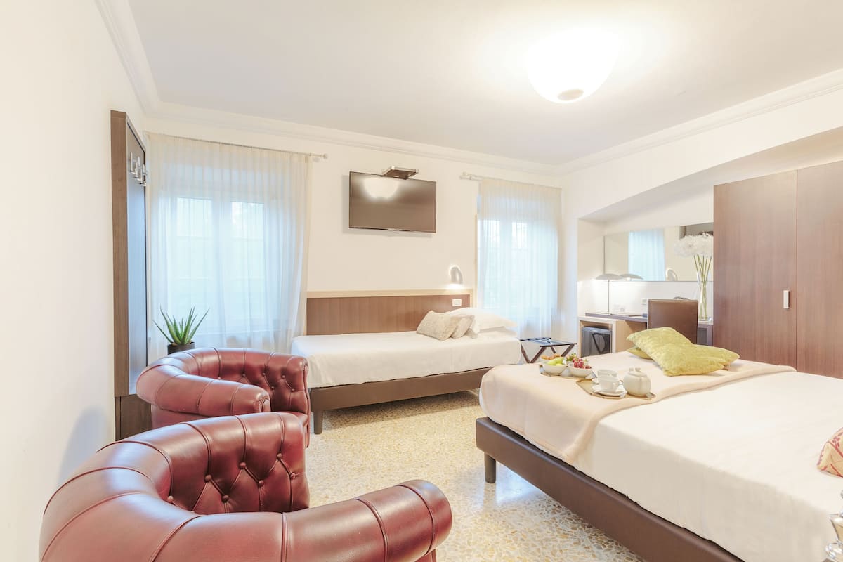 A hotel room with two beds, a red leather armchair, a bedside table with a lamp, and a large mirror.