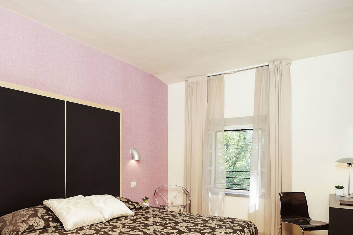 A bedroom with a bed, a wardrobe, a desk, a chair, a window with curtains, and a wall-mounted lamp.