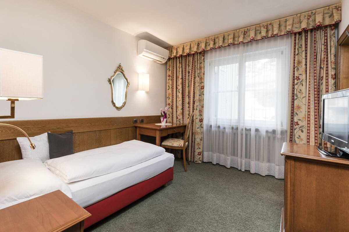 Habitación de hotel con cama, escritorio, silla, televisor, lámpara, espejo y ventana con cortinas.