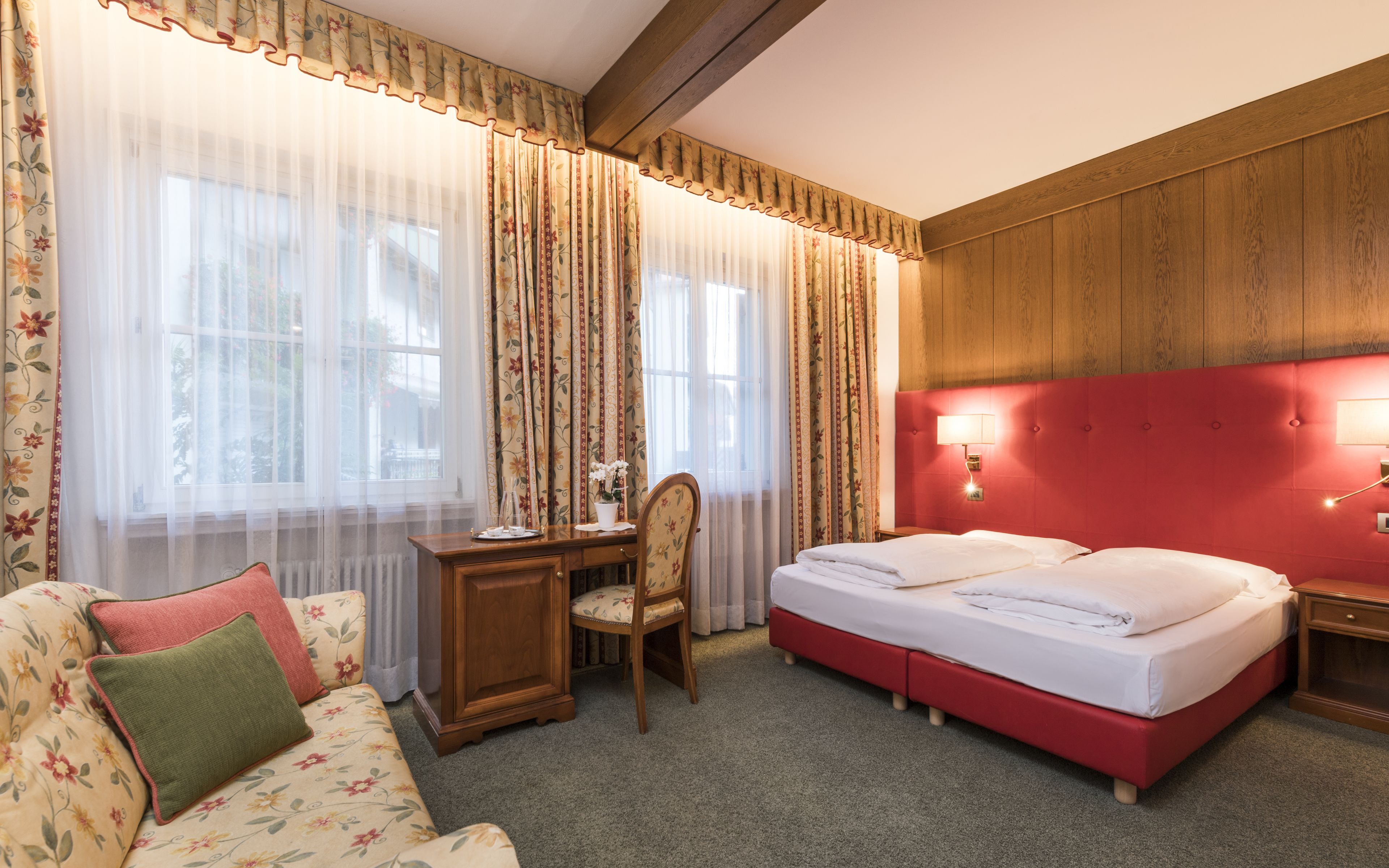 Ett hotellrum med en sänggavel i trä, ett skrivbord med en stol, en säng med två kuddar och en soffa med ett blommönster.