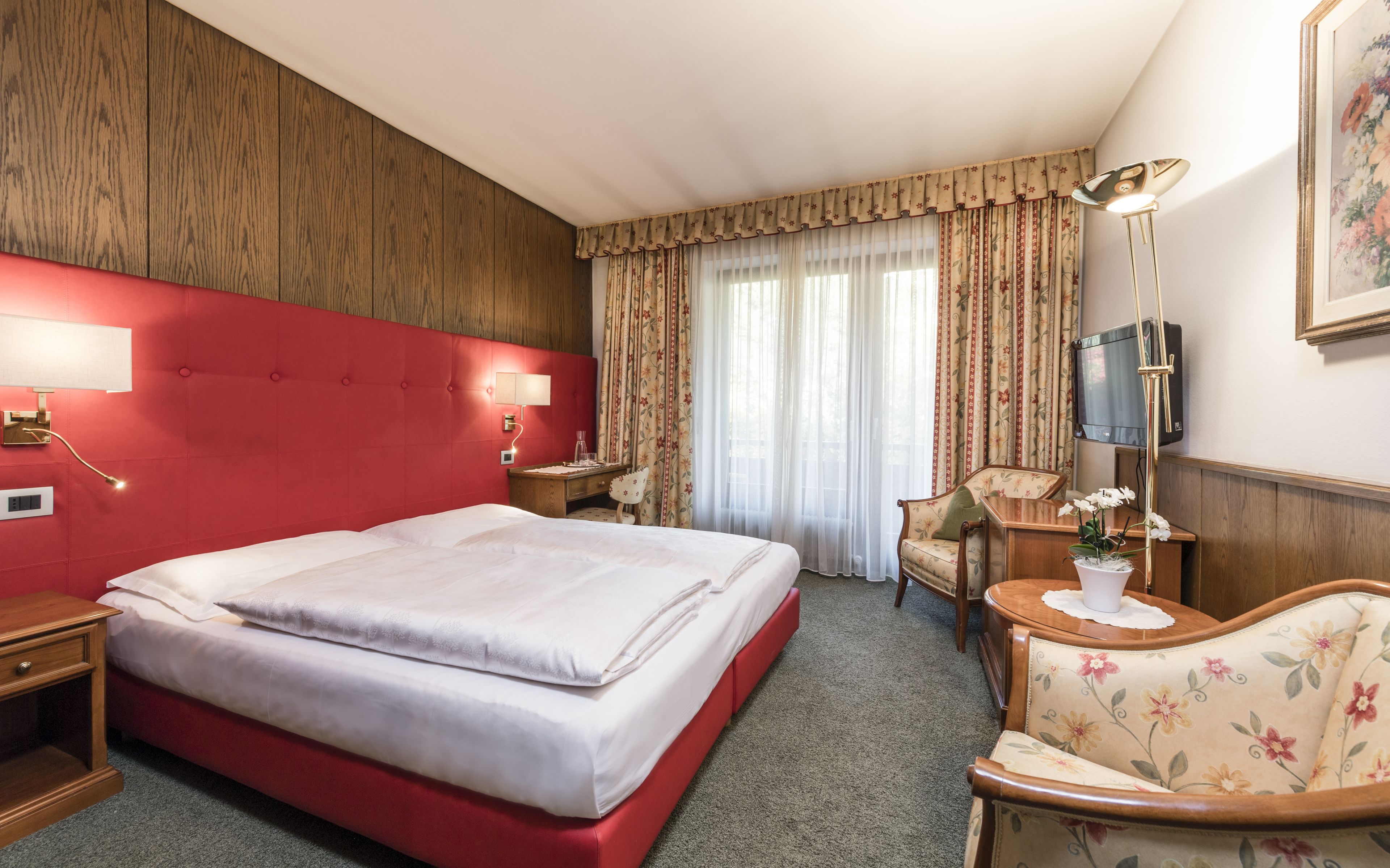 Une chambre d’hôtel avec une tête de lit rouge, un grand lit, une table de chevet en bois, un bureau avec une chaise, une lampe de bureau, un téléviseur et un fauteuil à motifs floraux.