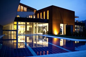 A modern building with a reflective pool, large windows, and a well-lit entrance. - GrünerBaum Hotels (Bressanone)