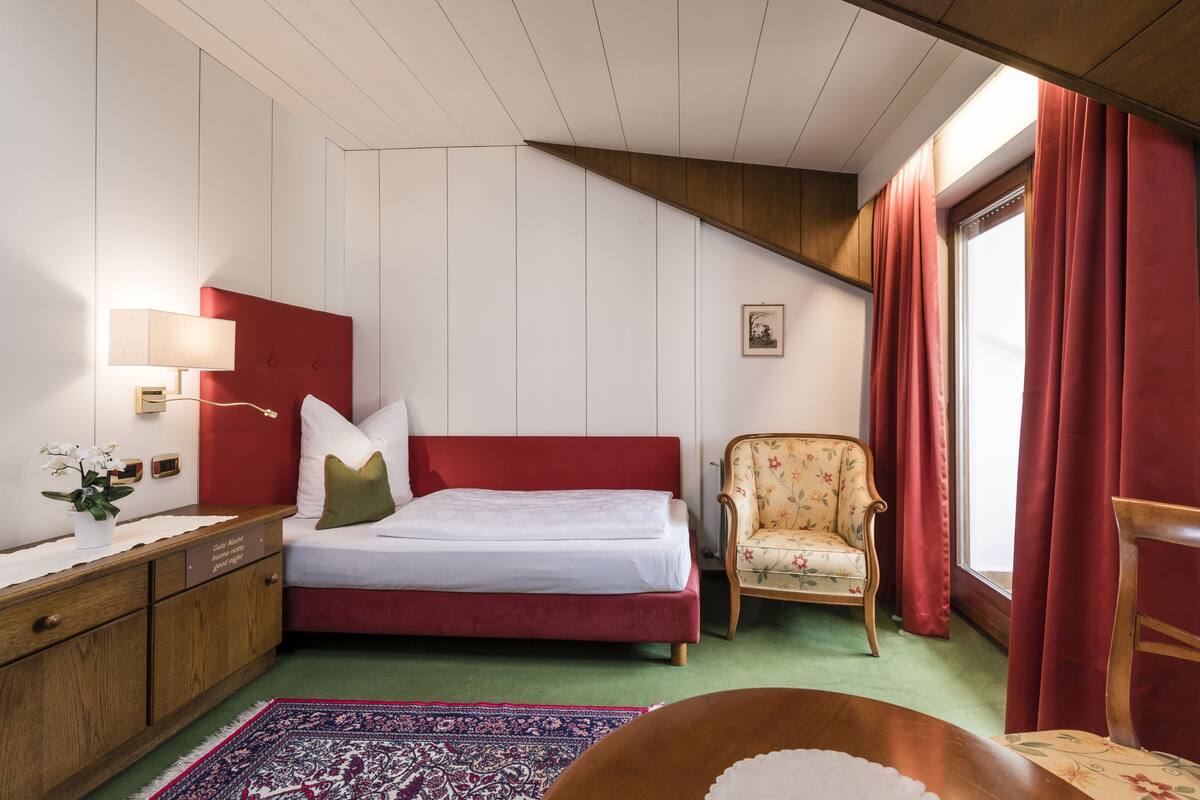 Un dormitorio con una cama roja, una mesita de noche de madera, un sillón estampado y vistas al exterior a través de una ventana con cortinas rojas.